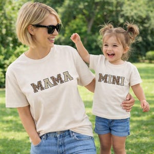 May include: Two cream-colored t-shirts with the words "MAMA" and "MINI" in a leopard print design. The shirts are worn by a woman and a young child, respectively. Both are smiling and wearing sunglasses and denim shorts.