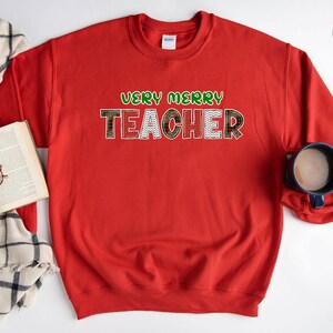 May include: Red sweatshirt with the text "Very Merry Teacher" in green and white letters with a red and green plaid pattern.