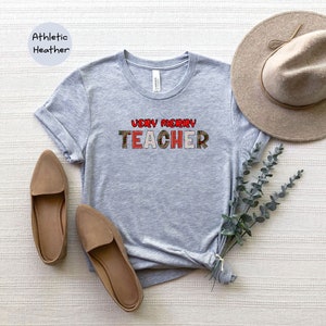 May include: A grey t-shirt with the text "Very Merry Teacher" in red, green and white plaid letters. The shirt is on a white surface with a brown hat and brown shoes.