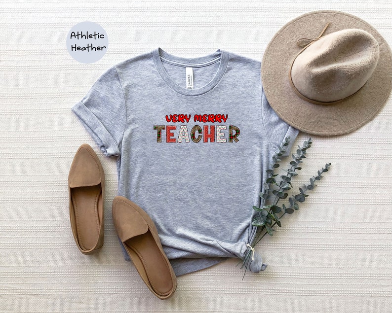 May include: A grey t-shirt with the text "Very Merry Teacher" in red, green and white plaid letters. The shirt is on a white surface with a brown hat and brown shoes.