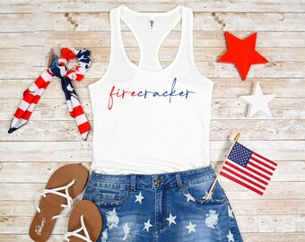May include: White tank top with the word "firecracker" in red and blue script, paired with denim shorts featuring white star patterns. Accessories include a red star, a white star, a small American flag, and sandals.