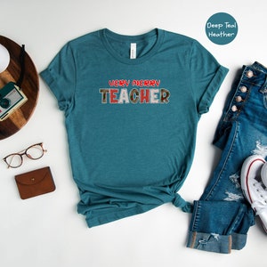 May include: A teal blue t-shirt with the text "VERY MERRY TEACHER" in red and white letters. The shirt is laid flat on a white background with a pair of jeans, a pair of white trainers, a brown wallet, a pair of glasses, and a vintage camera on a wooden tray.