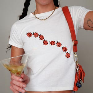 May include: White t-shirt featuring a curved line of red and black ladybug designs. The person is wearing a gold chain necklace and carrying a red handbag with a matching strap. A martini glass with olives is held.