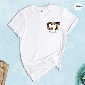 May include: White t-shirt with a leopard print "CT Tech" design on the chest. The text "tech" is written in a script font below the letters. The shirt is displayed on a light blue background with a pair of purple flip-flops and a straw hat.