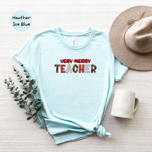 May include: Light blue t-shirt with the text "VERY MERRY TEACHER" in red and white letters with a Christmas-themed pattern.