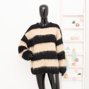 Può includere: Un maglione a righe nere e beige con scollo tondo. Il maglione è esposto su un manichino nero. Il maglione ha maniche lunghe e una consistenza soffice. Lo sfondo è bianco con una mensola decorativa.