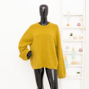 Può includere: Maglione giallo senape con girocollo e maniche lunghe. Il maglione è esposto su un manichino nero. Lo sfondo include una mensola bianca con oggetti decorativi.