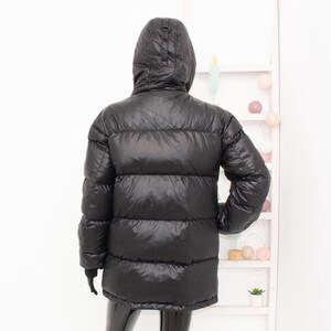 May include: Black puffer jacket with a hood. The jacket has a quilted design and a shiny finish. The jacket is a mid-length style, offering warmth and protection from the elements.