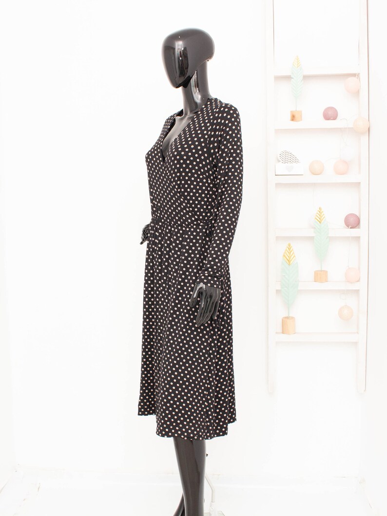 May include: A black midi dress with white polka dots. The dress has a V-neckline and long sleeves. It is cinched at the waist with a black belt.
