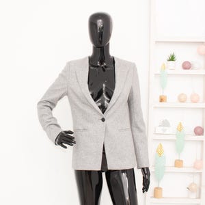 Pode incluir: Um blazer cinza-claro com um fecho de botão preto único ao centro, mangas compridas e dois bolsos horizontais com debrum na cintura. A peça de vestuário é exibida num manequim preto brilhante contra um fundo branco.