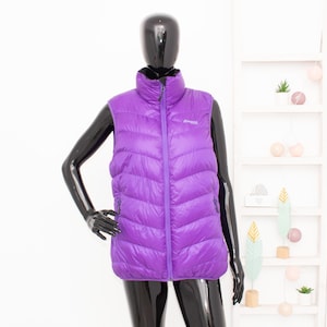 May include: A vibrant purple puffer vest with a high collar and a full-length zipper. The vest is displayed on a black mannequin. The vest has a quilted design and a small logo on the chest. The background is a white wall and a white shelf.