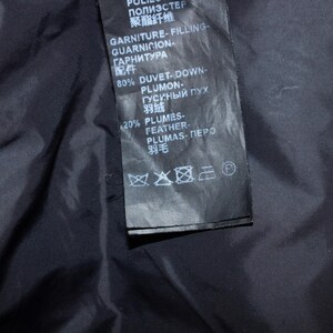 May include: Close-up of a black garment label with white text. The label includes information about the filling composition: 80% duvet-down and 20% feathers. The label also has care symbols.