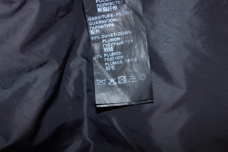 May include: Close-up of a black garment label with white text. The label includes information about the filling composition: 80% duvet-down and 20% feathers. The label also has care symbols.