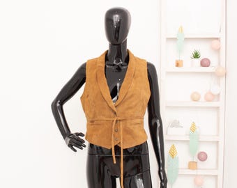 May include: A tan suede vest with a notched collar and a tie belt. The vest is displayed on a black mannequin. The vest has a button closure and two small pockets. The background is white with decorative shelves.