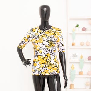Puede incluir: Una camiseta ajustada con cuello redondo y mangas hasta el codo, con un patrón denso y repetitivo de flores y follaje en amarillo, blanco y negro. La prenda se muestra en un torso de maniquí negro brillante sobre un fondo blanco.