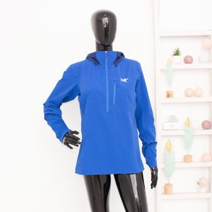 May include: A vibrant blue jacket with a hood and a quarter-zip closure. The jacket features a small logo on the chest and a streamlined design. The long-sleeved jacket is displayed on a mannequin.
