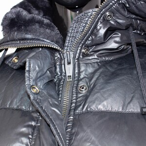 May include: Close-up of a black puffer jacket with a hood and a faux fur collar. The jacket has a zipper closure and snap details. The hood has a drawstring. The jacket is made of a shiny, water-resistant material.
