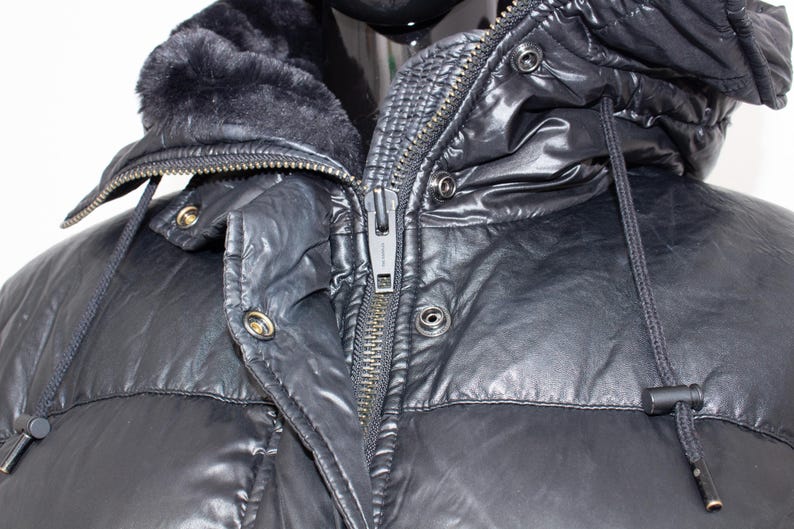 May include: Close-up of a black puffer jacket with a hood and a faux fur collar. The jacket has a zipper closure and snap details. The hood has a drawstring. The jacket is made of a shiny, water-resistant material.
