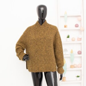 Può includere: Un maglione strutturato color senape con scollo a V è esposto su un manichino nero. Il maglione ha maniche lunghe e una vestibilità leggermente oversize. Il manichino è posizionato davanti a una mensola bianca con oggetti decorativi.