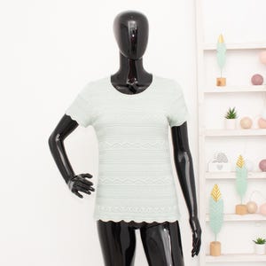 Könnte beinhalten: Hellgrünes Kurzarm-Shirt mit detailliertem Muster. Das Shirt hat einen gewellten Saum und wird auf einer schwarzen Schaufensterpuppe präsentiert. Das Design zeigt horizontale Bänder mit komplizierten Mustern.