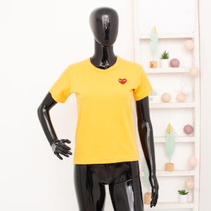 Op de afbeelding: Een gele t-shirt met korte mouwen en een rood hartlogo op de borst. Het shirt wordt getoond op een zwarte mannequin. De achtergrond is een witte muur met een decoratief rek.