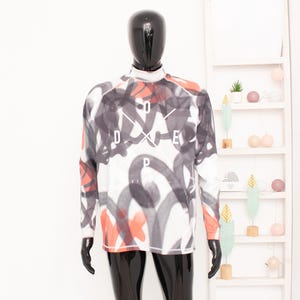 May include: A long-sleeved shirt with a high neckline, featuring a white base and a bold, abstract design in black, grey, and coral. The word "DOPE" is prominently displayed in white. The shirt is displayed on a mannequin.