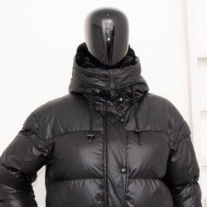 May include: A black puffer jacket with a high collar and a hood. The jacket has a shiny, quilted appearance and a front zipper closure. The mannequin is wearing the jacket in front of a white background.
