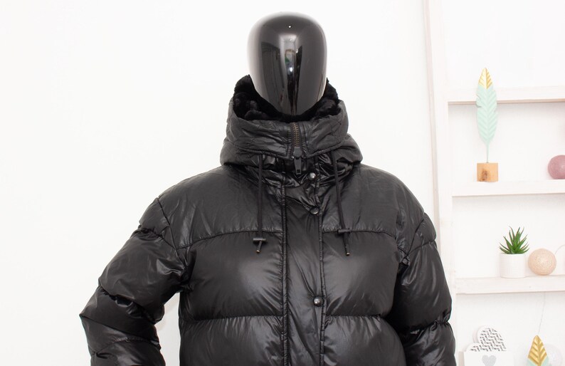 May include: A black puffer jacket with a high collar and a hood. The jacket has a shiny, quilted appearance and a front zipper closure. The mannequin is wearing the jacket in front of a white background.