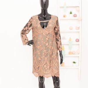 May include: A peach-colored lace dress with long sleeves and a knee-length hem. The dress features a floral pattern and a V-neckline with a decorative embellishment. The dress is displayed on a black mannequin.