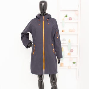 Może przedstawiać: Ciemnoszara kurtka z kapturem z pomarańczowymi zamkami i akcentami. Kurtka ma długi, opływowy krój, z wieloma kieszeniami i pełnej długości zamkiem błyskawicznym. Kurtka jest prezentowana na czarnym manekinie.