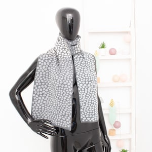 Pode incluir: Cachecol cinzento com um padrão de bolinhas brancas, drapeado sobre um manequim preto. O cachecol é feito de um material macio e tem uma borda. O manequim está em frente a uma prateleira branca com itens decorativos.