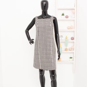 May include: A sleeveless shift dress in a black and white houndstooth pattern. The dress has a square neckline and is displayed on a black mannequin. The dress is knee-length and has a classic, timeless design.
