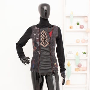 May include: A long-sleeved top on a mannequin. The top features a black turtleneck, with a patterned vest-like overlay in shades of gray, red, and brown. The overlay has decorative stitching and a red triangle accent. The sleeves and gloves are black.