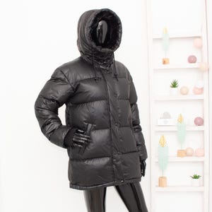 May include: A black puffer jacket with a hood, displayed on a mannequin. The jacket has a glossy finish and a quilted design. It features a high collar, a drawstring hood, and a button-up front. The mannequin is wearing black gloves.