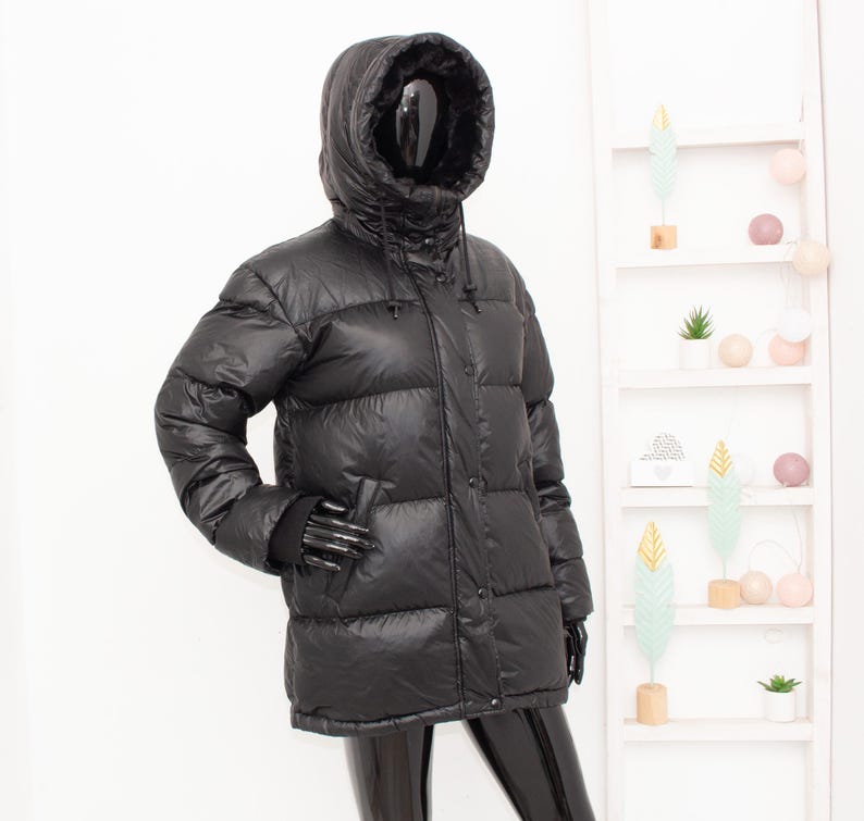 May include: A black puffer jacket with a hood, displayed on a mannequin. The jacket has a glossy finish and a quilted design. It features a high collar, a drawstring hood, and a button-up front. The mannequin is wearing black gloves.