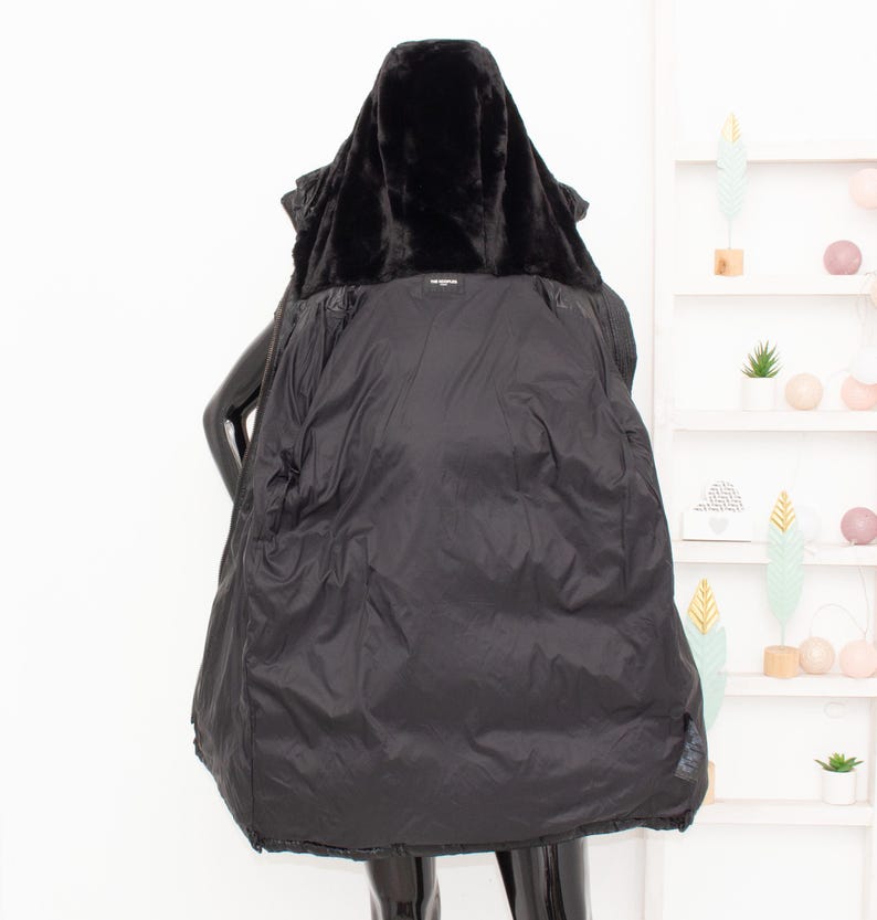 May include: A black winter coat with a faux fur-lined hood. The coat is open, revealing a quilted interior. The coat is displayed on a mannequin. The background includes a white shelf with decorative items.