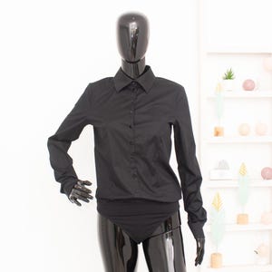 Può includere: Un body a camicia nero a maniche lunghe con colletto classico, abbottonatura frontale con bottoni neri coordinati e maniche aderenti. Il capo è esposto su un manichino nero lucido, che ne mette in risalto la vestibilità sartoriale.