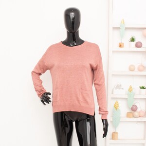 Puede incluir: Un suéter de manga larga y cuello redondo en color rosa apagado. El suéter se exhibe en un maniquí negro. El maniquí está colocado frente a un fondo blanco con una estantería decorativa.