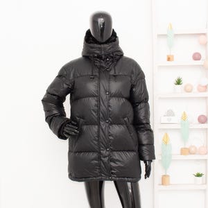 May include: A black puffer jacket with a hood, displayed on a mannequin. The jacket has a quilted design, a button-up front, and long sleeves. The mannequin is wearing black gloves. The background includes a white shelf with decorative items.