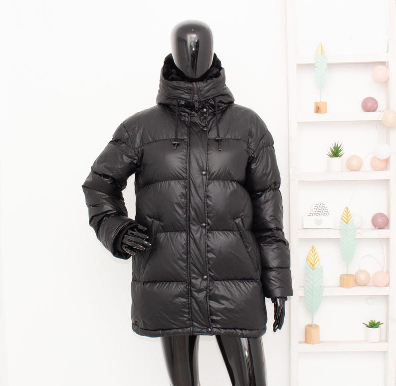May include: A black puffer jacket with a hood, displayed on a mannequin. The jacket has a quilted design, a button-up front, and long sleeves. The mannequin is wearing black gloves. The background includes a white shelf with decorative items.