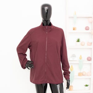 May include: A burgundy zip-up jacket with a high collar. The jacket has long sleeves and a full-length zipper. The garment is displayed on a black mannequin against a white background. The jacket appears to be made of a smooth, lightweight material.