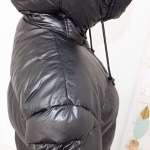 May include: Black puffer jacket with a hood and drawstring. The jacket has a shiny, quilted appearance. The hood is up, and the drawstring is visible. The jacket is a winter outerwear item.