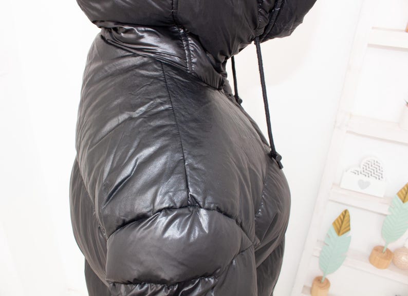 May include: Black puffer jacket with a hood and drawstring. The jacket has a shiny, quilted appearance. The hood is up, and the drawstring is visible. The jacket is a winter outerwear item.