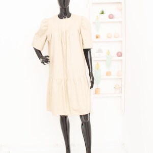 Peut inclure: Une robe beige arrivant au genou avec des manches courtes bouffantes, une encolure ronde et des plis verticaux sur la poitrine. Le vêtement présente une taille basse avec un panneau de jupe froncé qui ajoute du volume au bas de la robe.