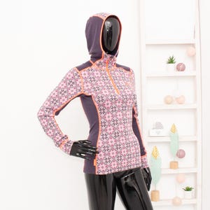 May include: A patterned athletic top with a hood, featuring a geometric design in pink and white, with orange detailing. The top has a quarter-zip and is displayed on a mannequin. The sleeves are long, and the design is sporty.