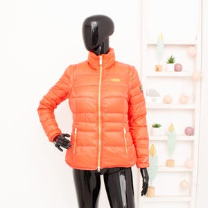 May include: A bright orange puffer jacket with a zip closure and a gold zip pull. The jacket has a stand-up collar and two pockets on the front.