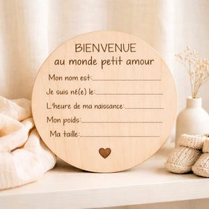 Peut inclure: Une plaque ronde en bois avec texte gravé en français : "BIENVENUE au monde petit amour". Elle comporte des lignes pour inscrire le nom, la date de naissance, l'heure, le poids et la taille du bébé. Un petit cœur est gravé en bas. Des chaussons de bébé sont visibles.