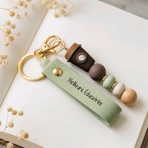 Peut inclure: Un porte-clés composé d'un anneau et d'un fermoir en métal doré, d'une lanière en similicuir vert sauge portant l'inscription "Meilleure Éducatrice" et d'une série de perles incluant une forme de tasse à café marron et des perles en bois.