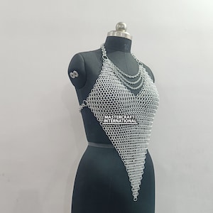 May include: Silver chainmail top with a V-neckline. The top is made of small, interlocking metal rings and has a triangular shape. The top is displayed on a black mannequin.