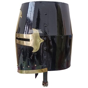 Op de afbeelding: Een zwarte, glanzende ridderhelm met messingkleurige accenten. De helm heeft een afgeronde bovenkant en een gezichtsbescherming met decoratieve messing elementen. De tekst "MASTER CRAFT INTERNATIONAL" is zichtbaar. Klinknagels bevestigen de metalen platen.